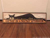 Image of a rectangular printed wooden plaque featuring a black cat lying amidst flowers with text 'CAT NAP ON A DOG DAY AFTERNOON'. Plaque resting on hardwood floor against white wall.