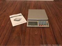 Photo showing the digital kitchen scale with control panel and LCD screen alongside its instruction manual, placed on a wooden floor.