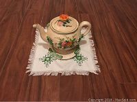Floral teapot with sculpted flower lid handle sitting on an embroidered white doily with green star patterns and fringe.