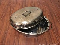Oval metal turkey roasting pan with matching lid and roasting rack inside, placed on wooden floor.
