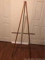 Full view of the vintage wooden tripod art easel standing upright against a wall on hardwood floor, showing its folding structure and horizontal support bar.