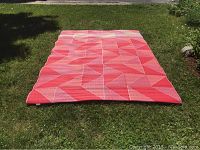 Outdoor rug laid flat on grass showing red faded side with geometric pattern.