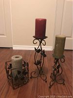 Three metal candle holders with leaf designs, each holding a different colored pillar candle. Sizes vary from small square base to tall curved base.