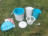 Photo showing the entire Greek yogurt maker set including the main unit, measuring pitchers, strainer, lid, and recipe/instruction booklets arranged on grass.