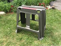Grey Rubbermaid plastic workbench outdoors on grass, showing the front and side with tool slots on top and built-in ruler.