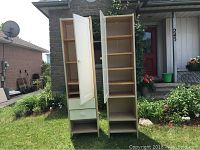 Two tall IKEA cabinets outside on grass, doors open showing interior shelves and drawers. Cabinets have cream-colored doors and light wood finish.