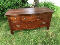 Antique low boy dresser placed outdoors showing front with three top drawers featuring round brass knobs and two larger bottom drawers with ornate brass handles, stained wood finish.
