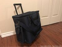 Side view of navy blue insulated portable fabric cooler showing telescoping handle, shoulder strap, and side pockets against closed door background.