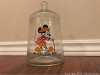Clear glass cookie jar with vintage Mickey Mouse design and removable lid, shown on a wooden floor against a wall.