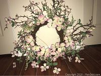 Photo of a circular artificial flower wreath featuring light pink and white flowers, green leaves, and small green berries on brown vine branches.