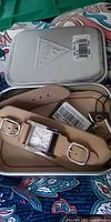 Watch in open Guess tin showing beige leather strap, silver-tone rectangular case, tags and barcode