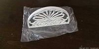 front view of white metal semicircular door stop with decorative cut-out fan design, resting on plastic bag