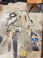 Group shot of all tools laid out on floor including saws, grease gun, plumber snake, pick, staple gun, ratchet, socket, roller frames, bulb changer pole