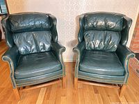 Front view of both dark green leather wingback chairs showing nailhead trim and wood legs