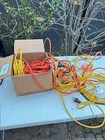 Box containing coiled yellow and orange extension cords, additional cords on table with plug visible