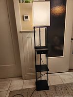 Full view of black floor lamp with white shade, drawer and shelves