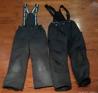 Two pairs black snow bib pants side by side showing suspender straps and overall wear