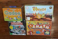 Group shot of four game boxes: Cadoo tin, Say Cheese, U-Build Monopoly, Smath