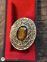 Front view of gold-tone oval pendant compact with amber cabochon and raised pattern on red insert
