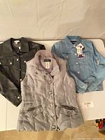 All three outerwear items laid flat on table: black faux leather jacket, grey quilted vest, light blue denim jacket with tag