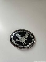 Front of oval belt buckle showing silver eagle on black background with floral border and rope edge