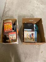 Two boxes filled with assorted books of various genres and sizes on concrete floor