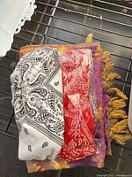 Stack showing white paisley bandana, red bandana in plastic, and purple-gold fringe wrap underneath