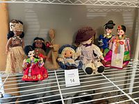 Shelf view showing seven dolls: hula dancer, Indigenous doll, burlap figure, small rag doll, large rag doll, two Korean costume dolls