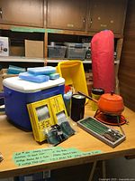 Overall view of cooler, tent bag, stove, flashlights, basin, skewers, forks, thermos, clothespins