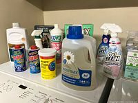 Multiple cleaning products on washing machine including eco max detergent jug, Drano, OxyPro, OxiClean sprays