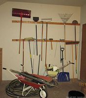 Overall view showing wheelbarrow, hose, spreader, snow shovel and multiple long-handled tools mounted on wall.