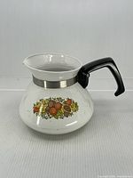 Full view of white enamel Corning Ware coffee carafe with black handle and vegetable motif