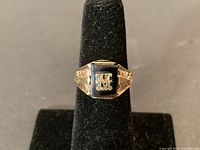 Front view showing black stone with raised "M" and gold setting