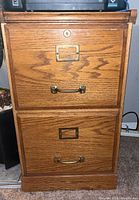Front view of oak two-drawer file cabinet showing brass-tone pulls, label frames and keyhole