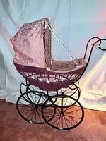 Left side view of wicker doll carriage showing metal frame, wheels, wicker basket, pink canopy
