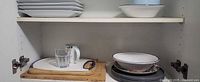 Cabinet shelf with stack of rectangular plates, serving bowl, blue bowl, mug, glass and cutting boards