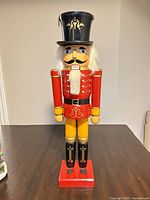 Front view of painted wooden nutcracker showing red uniform, black hat, white hair/beard, missing boot fronts