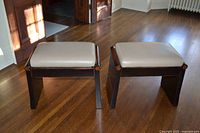 Two matching cherry wood base footstools with beige leather tops on hardwood floor