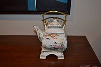Front view of porcelain teapot on stand showing floral and bird motif, metal handle visible