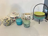 Group shot of all items: two stacking teapot sets, small pitcher, two lidded cups, metal stand with seven coloured plates