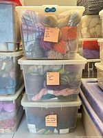 Three stacked clear plastic tubs filled with assorted skeins and hanks of yarn, sticky notes showing counts