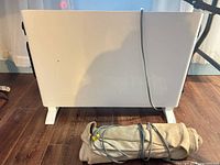 White DeLonghi panel heater standing on feet; beige heating pad rolled in front, cords visible