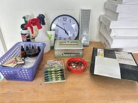 Full lot overview on tabletop showing clock, spray bottles, knit kit tin, coins dish, basket with various bottles and matchboxes, stack of postcards, black parcel box, stack of white boxes