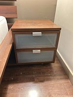 Nightstand front view showing wood veneer case, two frosted drawer fronts, caster base