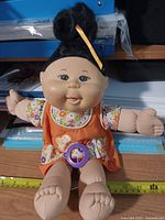 Front view of doll seated on desk showing dress, limbs, pacifier, measuring tape