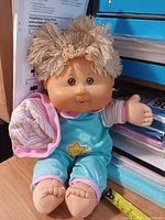 Front view of 14-inch Cabbage Patch Kids girl doll seated on desk showing blue sleeper, striped sleeves, blonde yarn hair, brown eyes