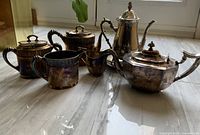 Group view of six of the eight silver-tone tea service pieces on marble floor