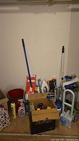 Overview of floor showing mop bucket, broom, spray mop, iron, cleaners in box