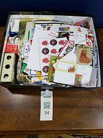 Top-down view of shallow box containing mixed Christmas gift tags, stickers, small cards, envelopes, ribbons and string