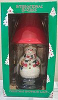 Front view of boxed International Bazaar Christmas Snowman Lamp showing snowman figure and red shade through window